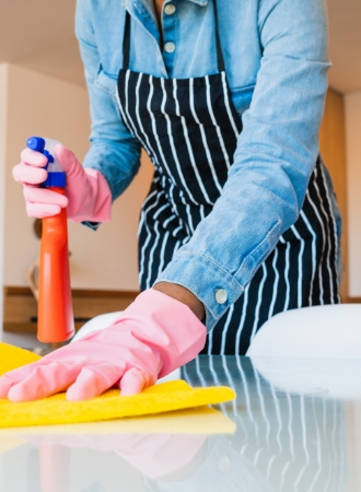 Person wearing pink gloves and a striped apron is cleaning a glass table with a yellow cloth, holding an orange spray bottle. The scene conveys tidiness.