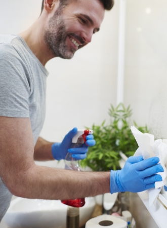 A smiling man in a gray shirt and blue gloves cleans a bathroom mirror with a spray bottle and paper towels. The scene is bright and tidy, with a plant visible in the background.