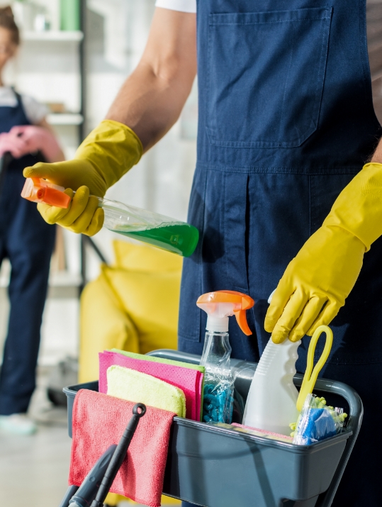 Person in a blue apron and yellow gloves holding a spray bottle with green liquid over a cleaning cart. Focus on cleaning supplies, suggesting domestic work.